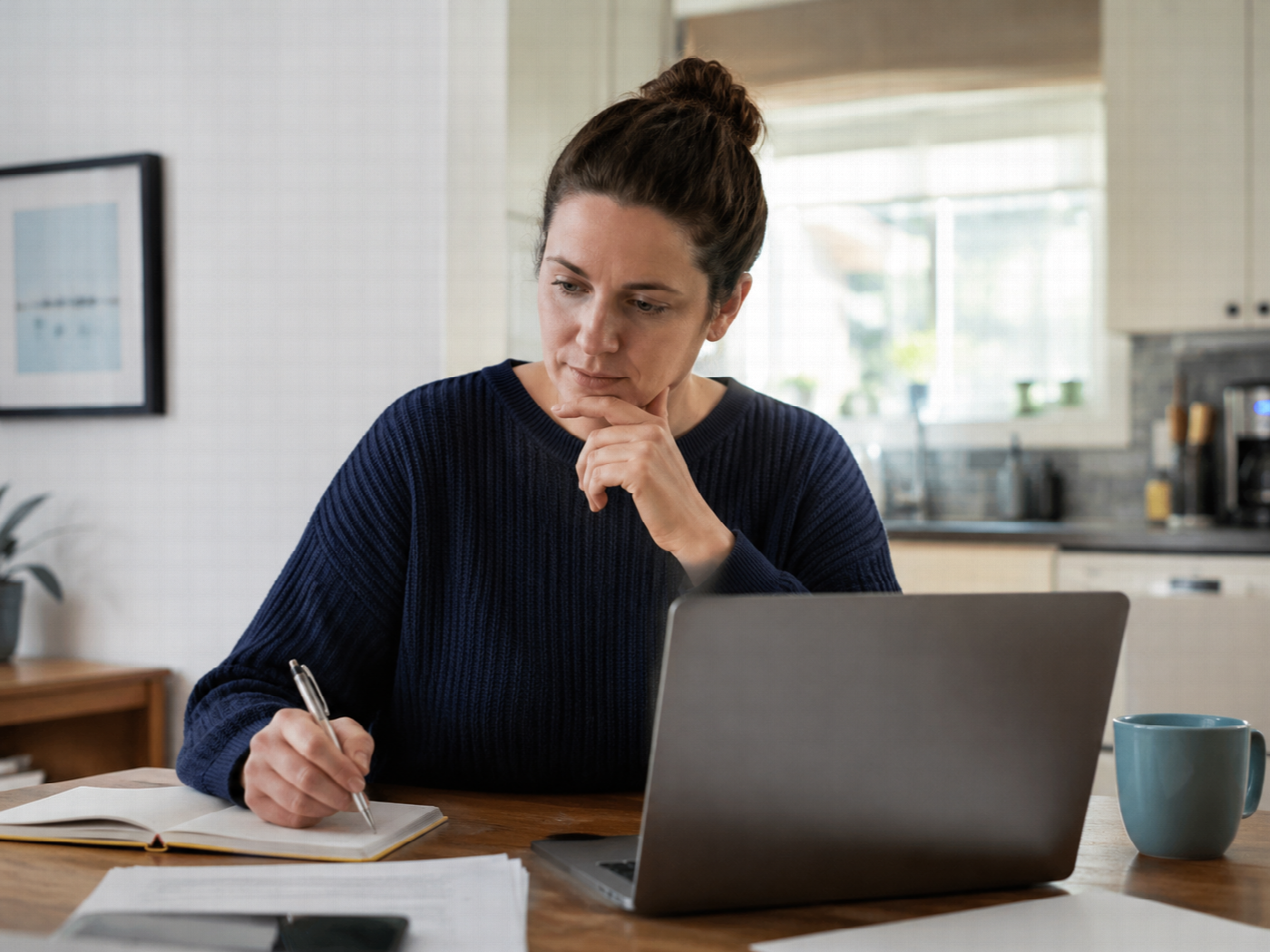 A person at a kitchen table checking a provider on a laptop with a notebook and pen beside them.